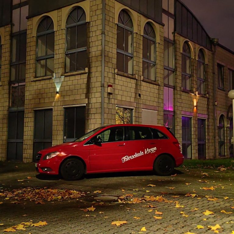 Mercedes B 180 CDI Rotes Auto parkt vor einem modernen Gebäude mit bunten Lichtakzenten.